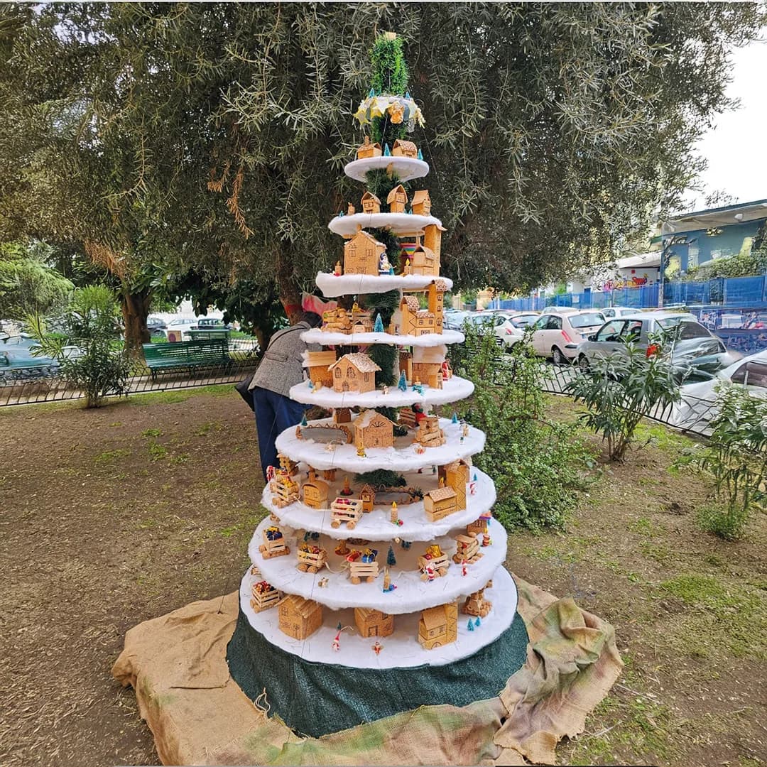 L’albero di Natale riciclato: una torre di ripiani bianchi con casette di cartone e sughero, sotto un ulivo